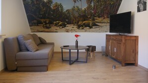 A living room with a sofa, a coffee table with a vase, a TV on a wooden cabinet, and a mural of dinosaurs on the wall. - Vacation cottage Baarsen 30 (Bad Pyrmont)
