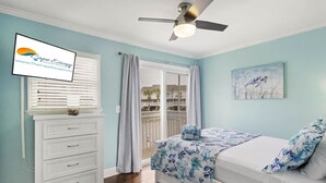 Quarto com cama, ventilador de teto, janela com persianas, penteadeira branca e vista para uma varanda.