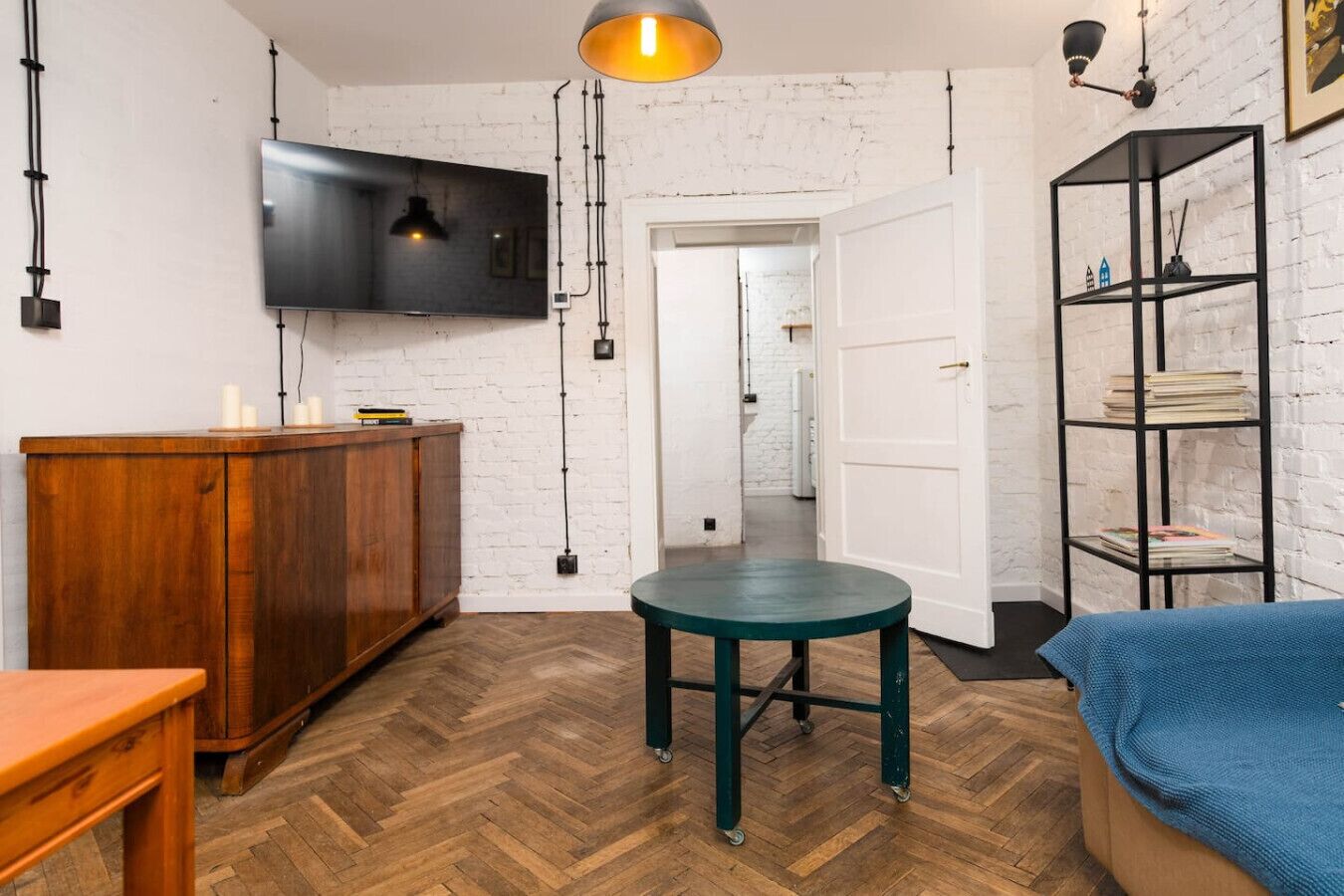 Una habitación con piso de madera en patrón de espiga, un mueble de madera, una mesa turquesa, un televisor negro montado en una pared de ladrillo blanco y una estantería de metal negro.