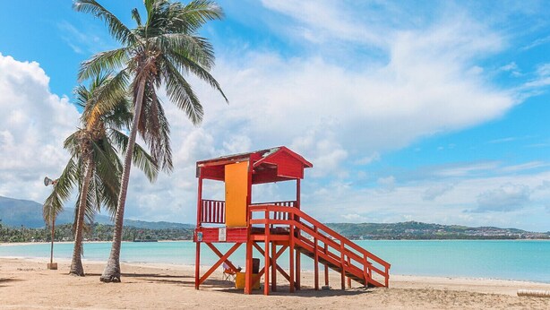 A beach with a red lifeguard tower, palm trees, and clear blue water. - Ocean View in a central apartment w/ pool and gym (Fajardo)