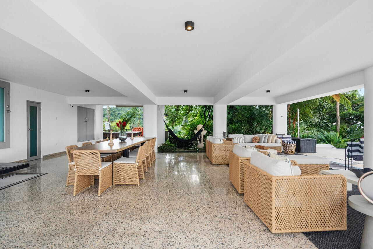 A spacious indoor area with a dining table, wicker furniture, and a view of lush greenery outside.