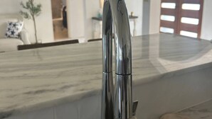 Uma cozinha moderna com torneira de aço inoxidável, bancada de mármore e vista para a sala de estar, que inclui um sofá e uma planta.