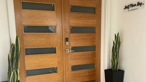 Uma porta dupla de madeira com painéis de vidro e fechadura eletrônica, ladeada por vasos de plantas, com um tapete listrado à frente.