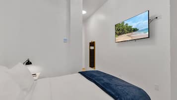 A modern hotel room with a bed, a TV mounted on the wall displaying a beach scene, a bedside table with a lamp, and a door leading to another room.