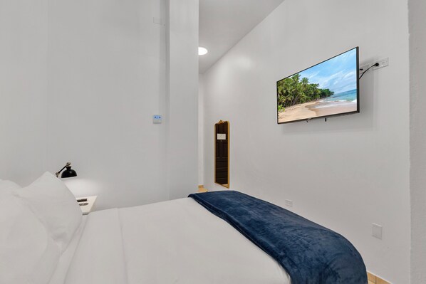 A modern hotel room with a bed, a TV mounted on the wall displaying a beach scene, a bedside table with a lamp, and a door leading to another room. - Armas Hotel (San Juan)
