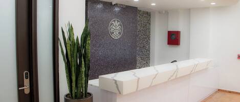 Un área de recepción con mostrador de mármol, una planta en maceta, un panel decorativo y un detector de incendios.