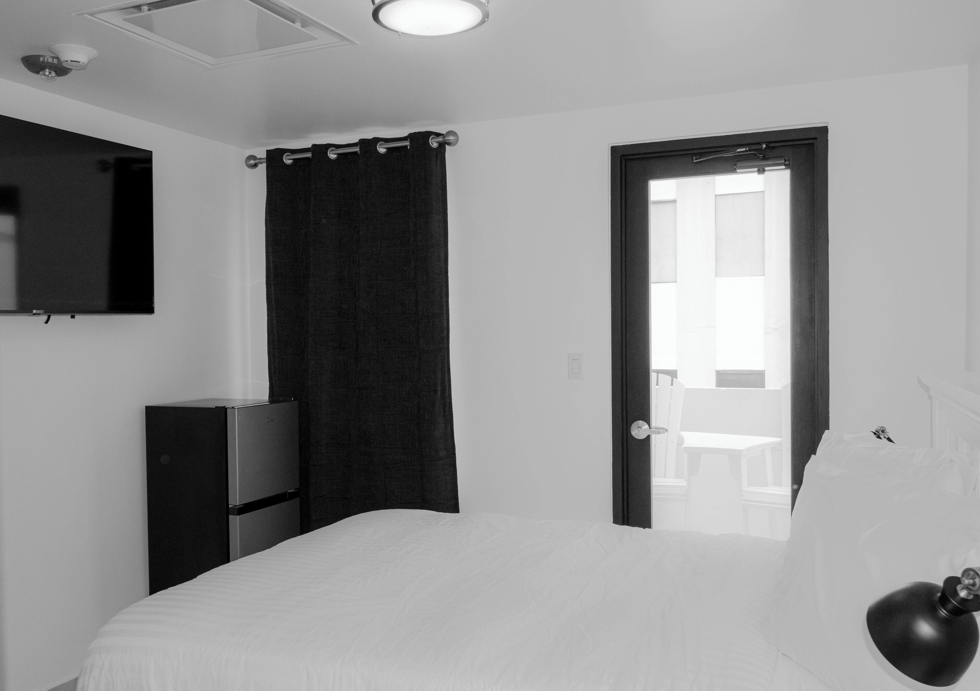 Habitación de hotel minimalista con una cama, un televisor de pantalla plana en la pared, una cortina oscura y una puerta que da a un balcón.