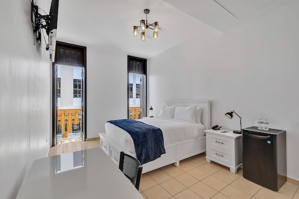 A modern hotel room with a bed, bedside table, desk, and a small refrigerator.