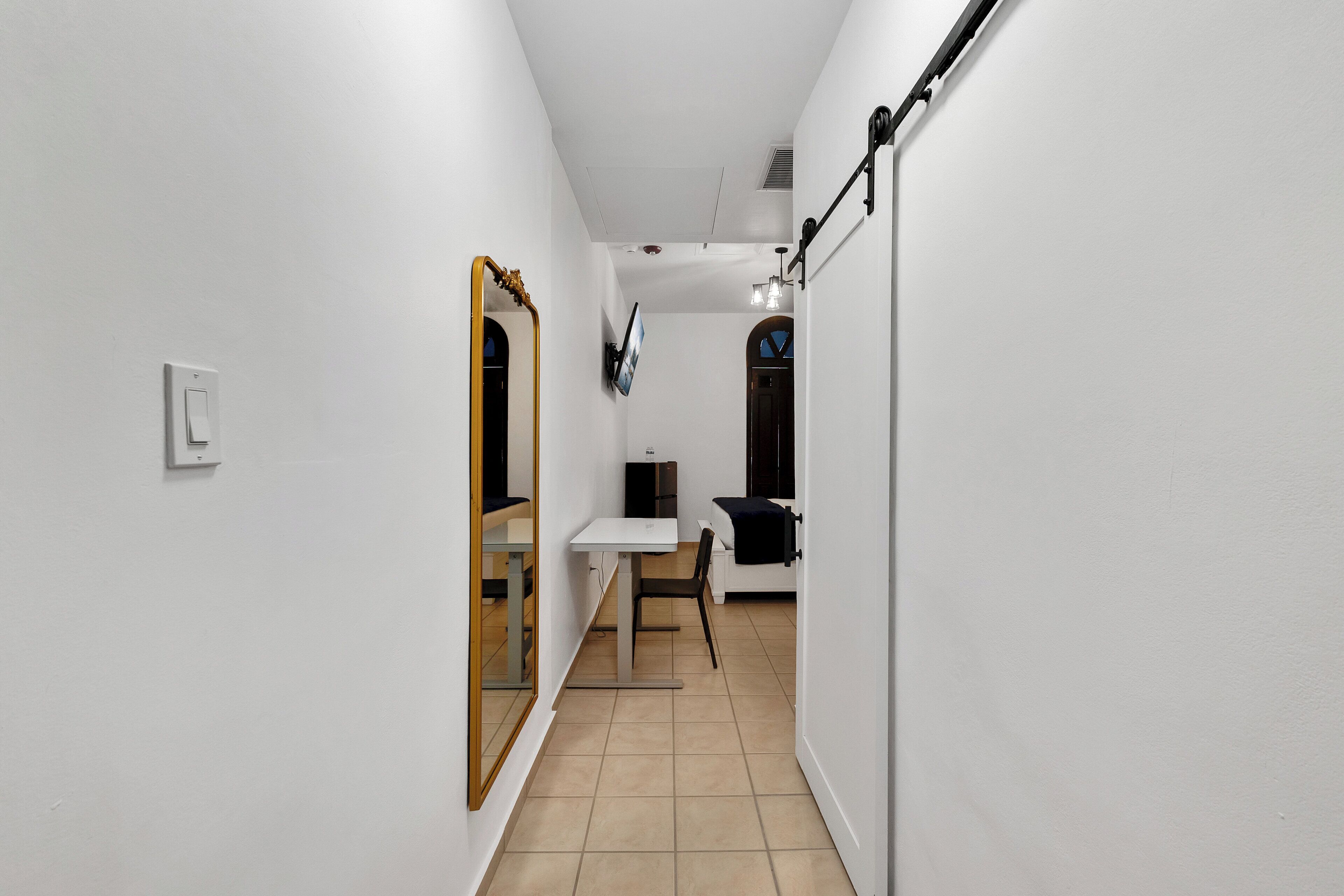 A narrow hallway with a white wall, a wooden-framed mirror, a white table with a chair, and a television mounted on the wall.