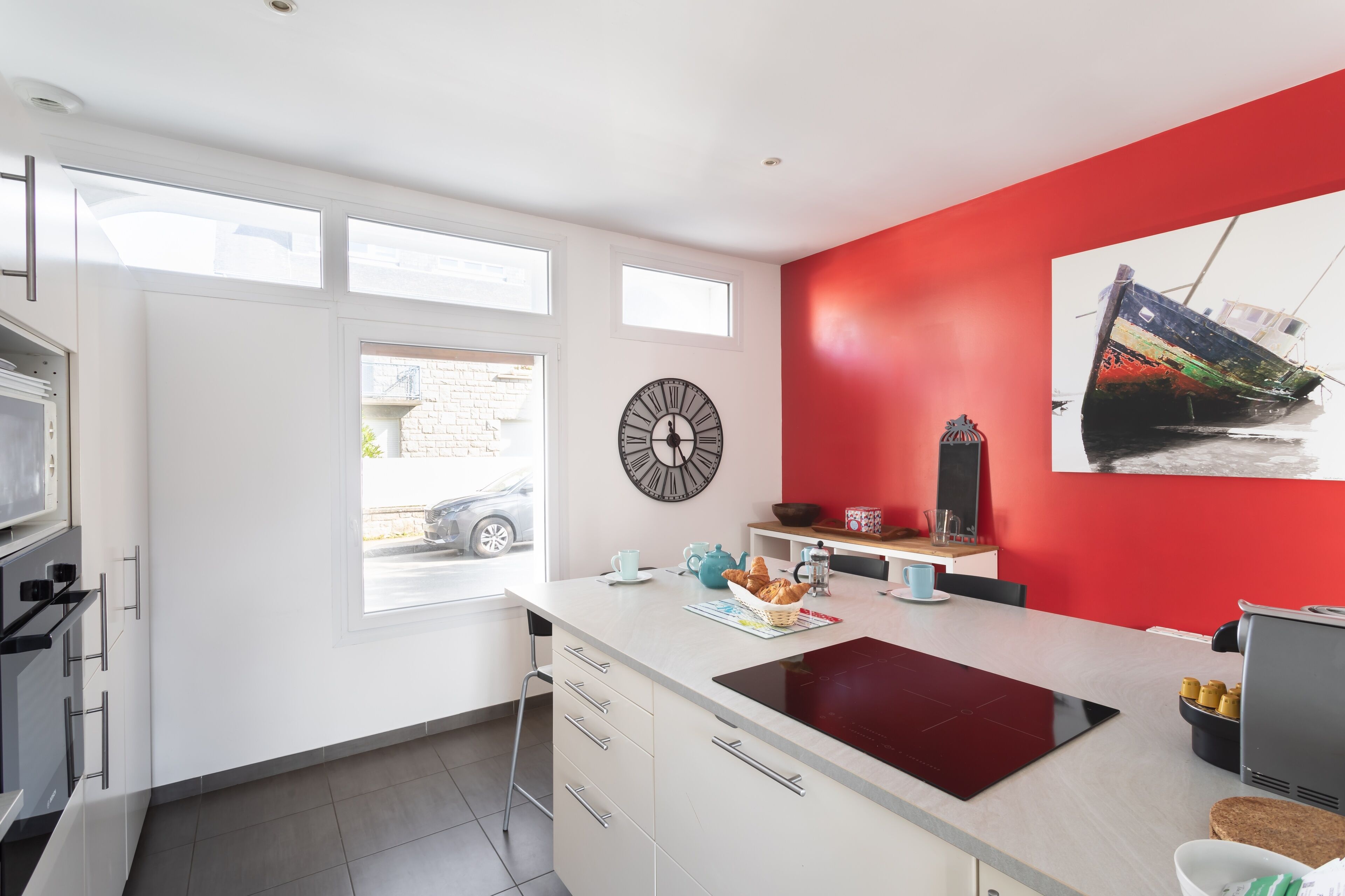 A modern kitchen with white cabinets, a red accent wall, a large window, and a clock.