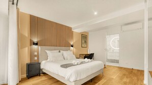 Een moderne slaapkamer met een houten lattenkap, een bed met witte lakens, een nachtkastje met een lamp, en uitzicht op een badkamer met een ronde spiegel.