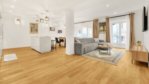 Een ruime woonkamer met een grijze bank, houten vloeren, een eettafel met stoelen, een keukenhoek en grote ramen met gordijnen.
