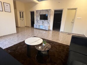 A modern living room with a marble floor, a round table with a white marble top, a dark sofa, and a flat-screen TV mounted on a marble wall. - Eraya Abode (Mysore)
