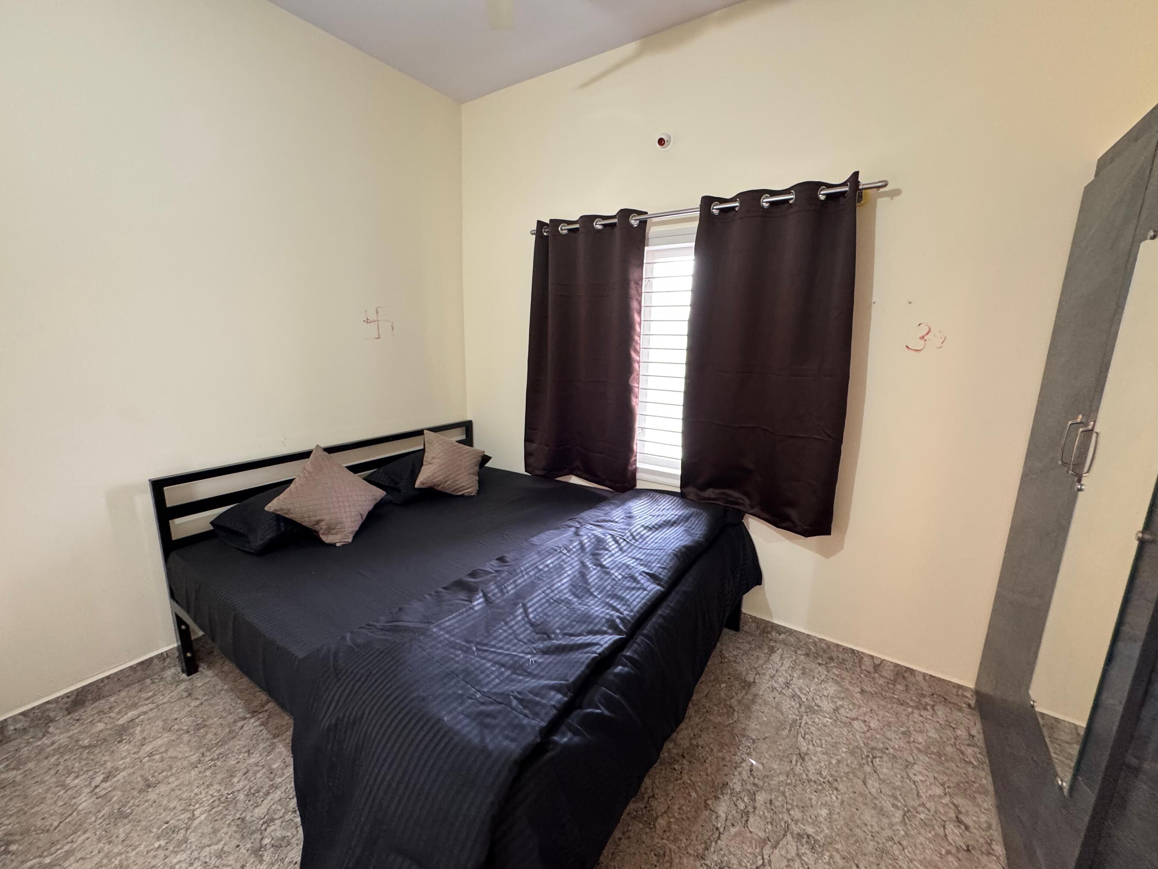 Un lit simple avec un matelas et des oreillers noirs, une fenêtre avec des rideaux foncés et une porte sur laquelle est inscrit le numéro « 3 ».