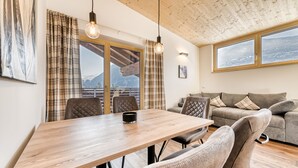 A modern living room with a wooden dining table, comfortable seating, and large windows offering a mountain view. - Alpenblick Appartements - 3-zimmer Dachgeschoss (Schruns)