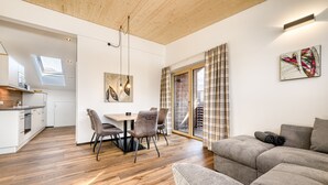 A modern living room with a sofa, a dining area with chairs, a kitchen, and a large window with curtains. - Alpenblick Appartements - 3-zimmer Dachgeschoss (Schruns)