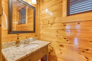 A wooden cabin bathroom with a granite countertop sink, a mirror, and a window with blinds.
