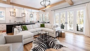 Een ruime woonkamer met een grote witte modulaire bank, een zebra vloerkleed en een moderne keuken met witte kasten en hangende lampen.