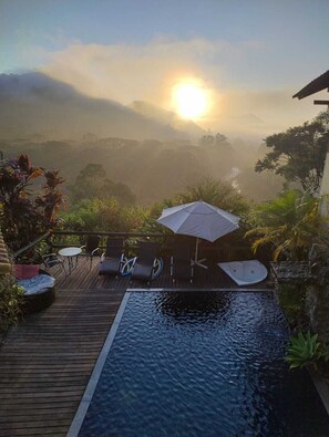 A wooden deck with a hot tub, lounge chairs, and an umbrella, overlooking a misty landscape with a sunrise. - Pousada Bella Vista (Itatiaia)