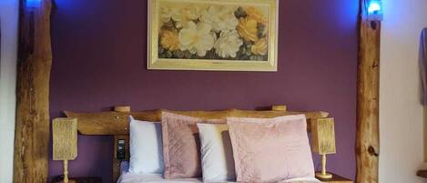 A bedroom with a wooden headboard, a large bed with a patterned bedspread, two bedside lamps, and a wall-mounted painting.