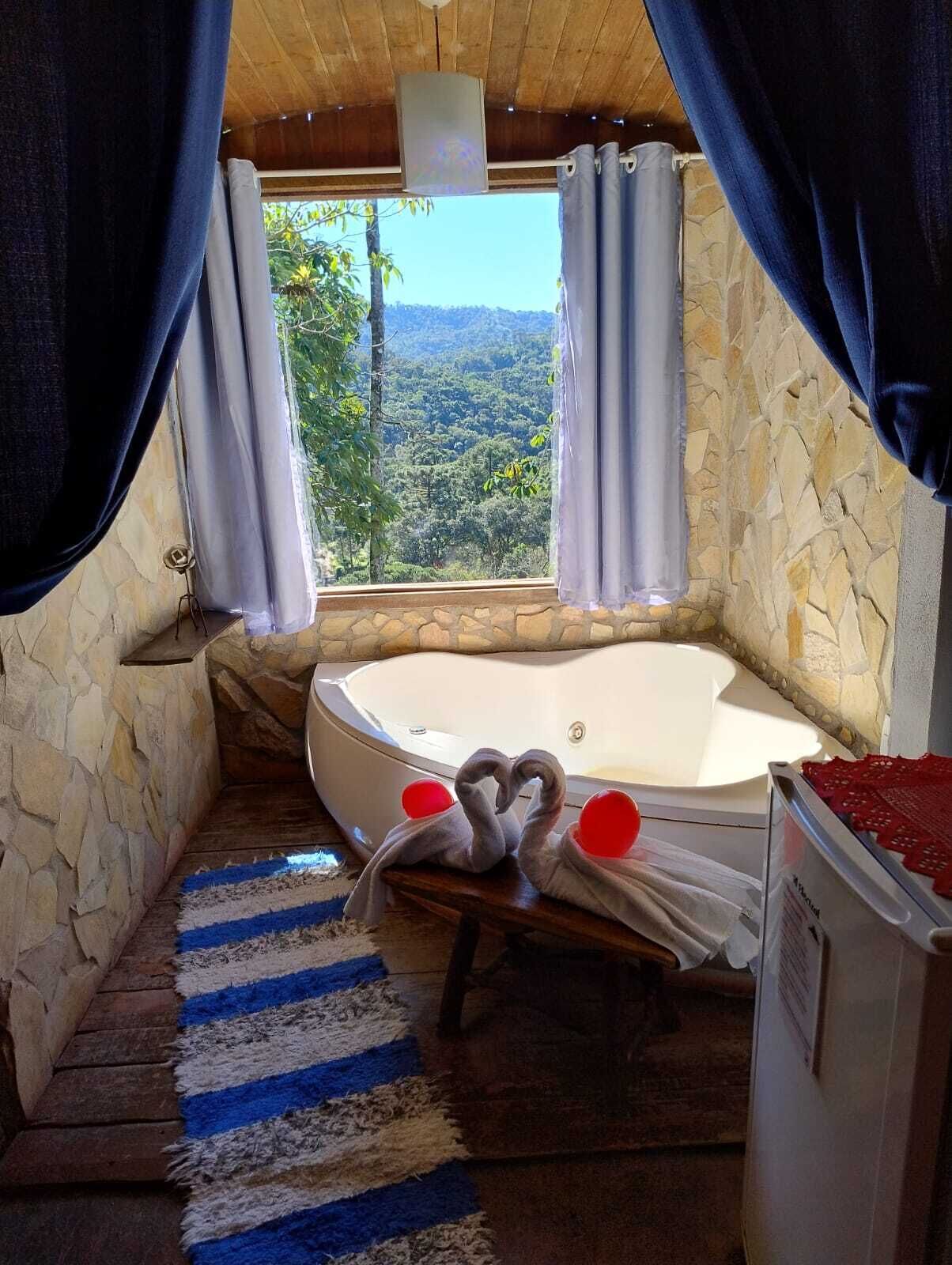 A room with a stone wall, a bathtub, a striped rug, and a view of a forest.