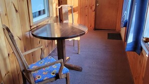 Interno di una baita in legno con un tavolo rotondo, due sedie, una porta in legno e una finestra con tende.