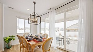 Een eetruimte met een houten tafel voor vier personen, omringd door stoelen, een hanglamp en uitzicht naar buiten door grote ramen.