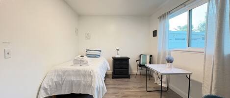 Un dormitori amb un llit, escriptori, cadira, prestatge i finestra amb cortines.
