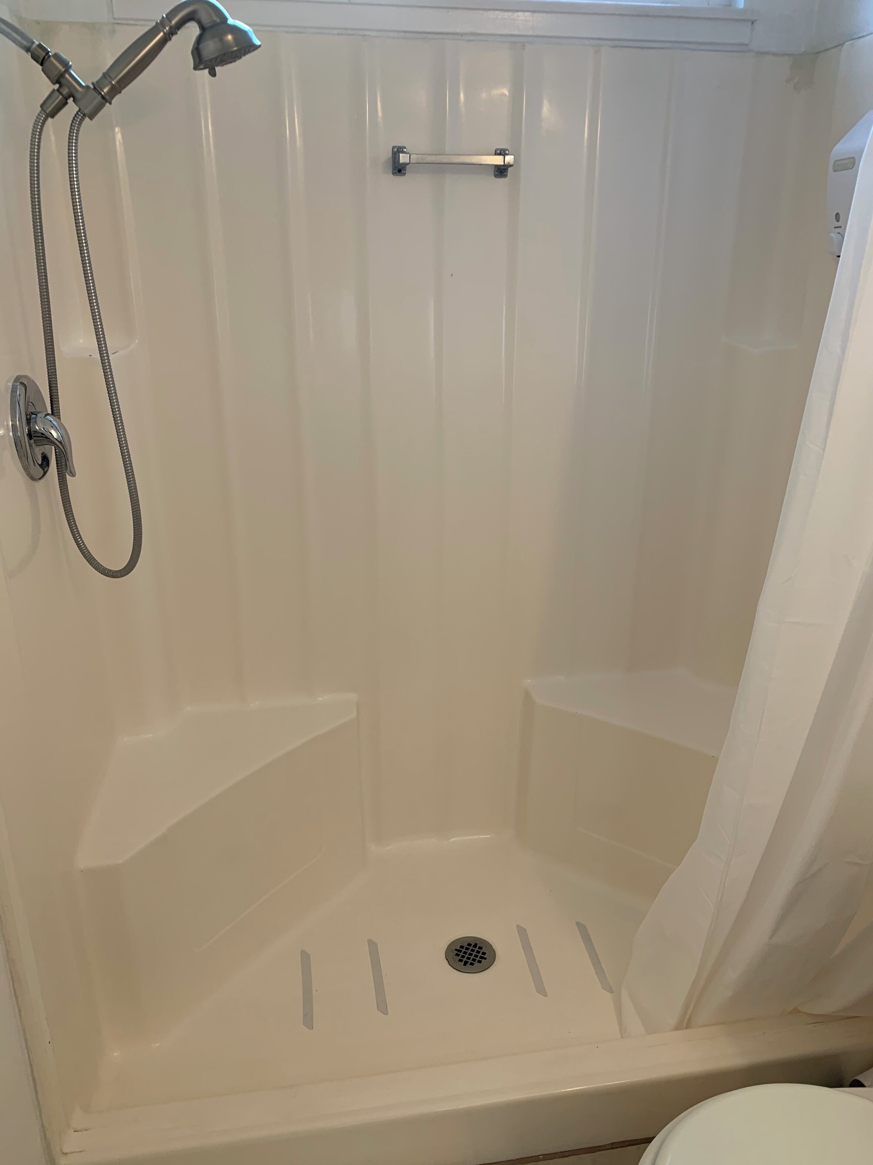 A white shower enclosure with a handheld showerhead and a fixed showerhead, a drain in the center, and a towel rack on the wall.