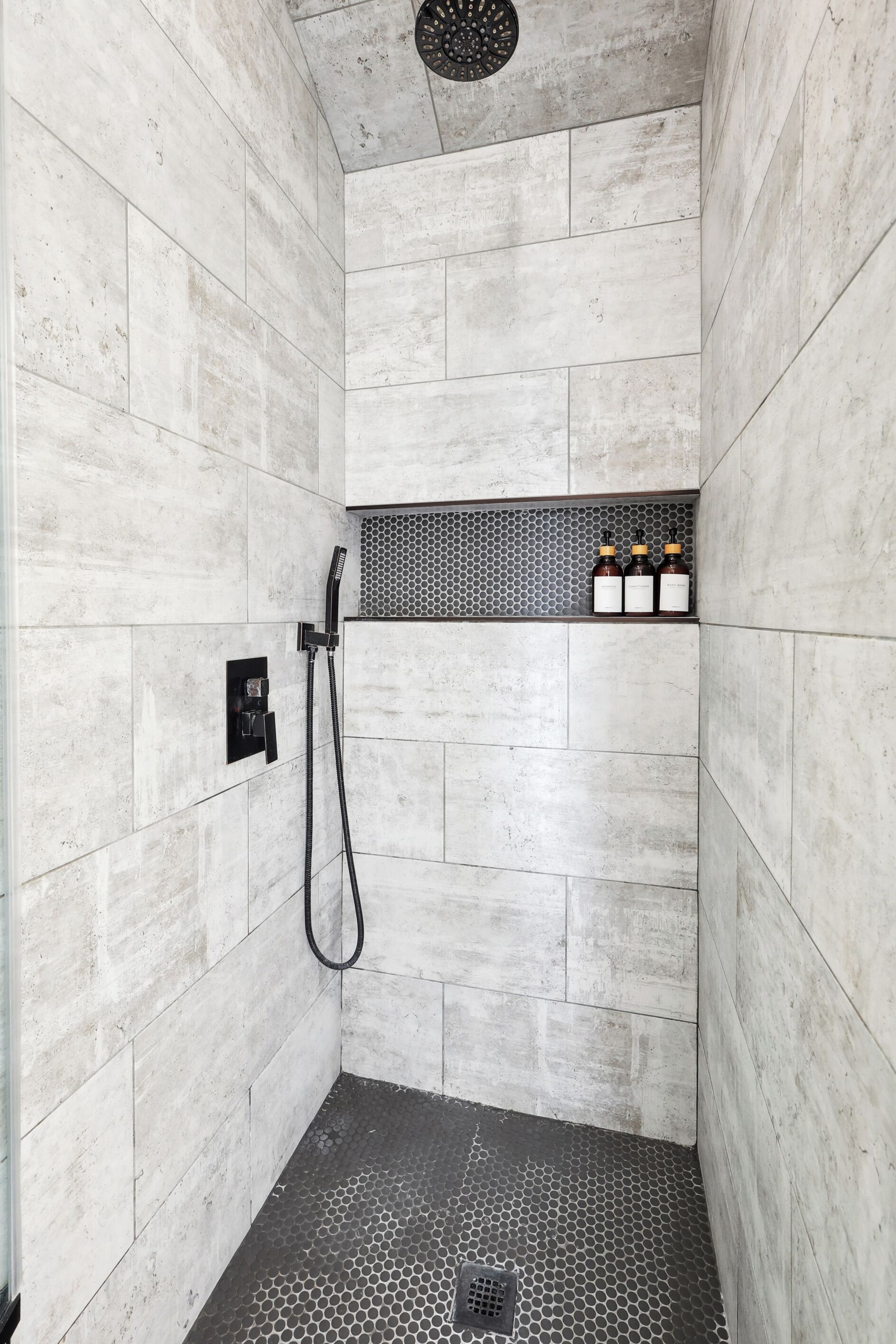 A shower area with a tiled floor, a showerhead, and a shelf with bottles.