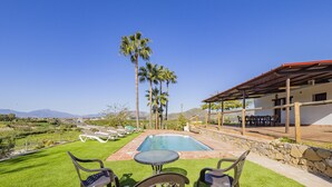 Zona de piscina con vistas a montañas, un porche cubierto con mobiliario exterior y un césped bien cuidado.