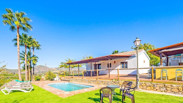 Zona de piscina con solárium de piedra, terraza cubierta con sillas y vistas a montañas al fondo.