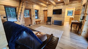 A cozy cabin interior with a wooden floor, a sofa, a bench, a television, and a window overlooking a forest. -  Play House Private Ozone Hot Tub, w/ BEAUTIFUL views. Quaint, Cozy, & Peaceful. (Sandpoint)