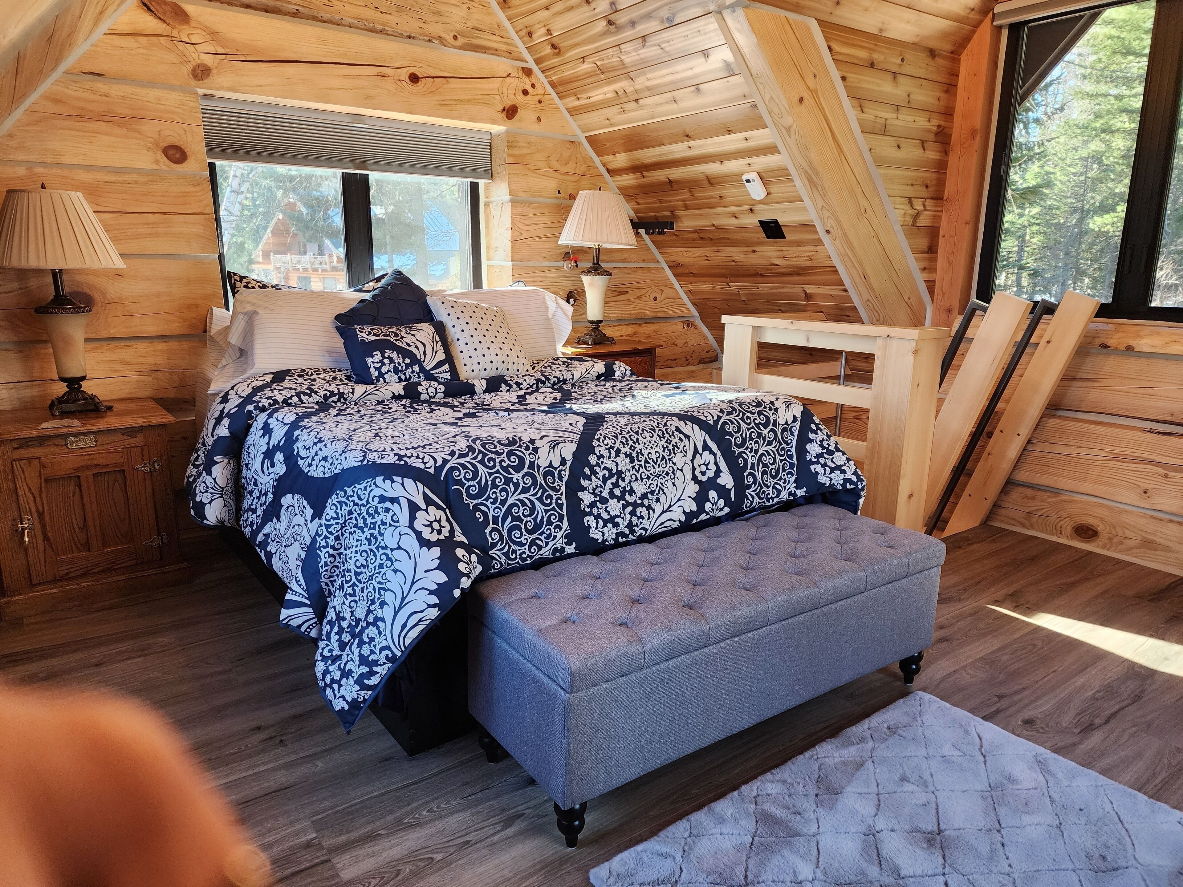 Un dormitorio acogedor con techo de madera, una cama grande con colcha estampada, dos mesitas de noche con lámparas y vistas al exterior a través de una ventana.