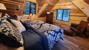 A cozy attic bedroom with a wooden sloped ceiling, a bed with patterned bedding, a dresser, and two windows offering views of the surrounding forest. -  Play House Private Ozone Hot Tub, w/ BEAUTIFUL views. Quaint, Cozy, & Peaceful. (Sandpoint)