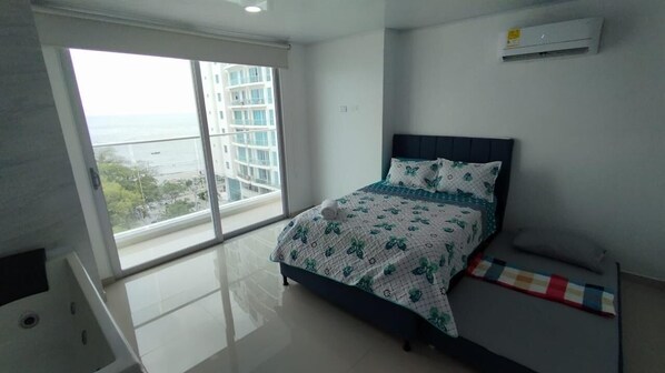 A bedroom with a bed, a window with a view of buildings and a balcony, and an air conditioning unit on the wall.