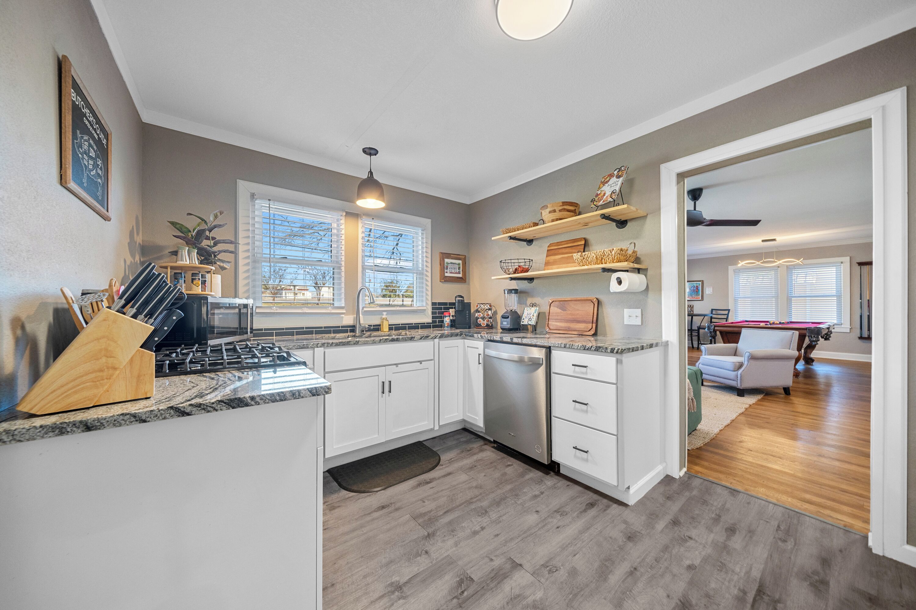 A modern kitchen with white cabinets, a stainless steel dishwasher, and a granite countertop. There is a dining area visible through an open doorway.