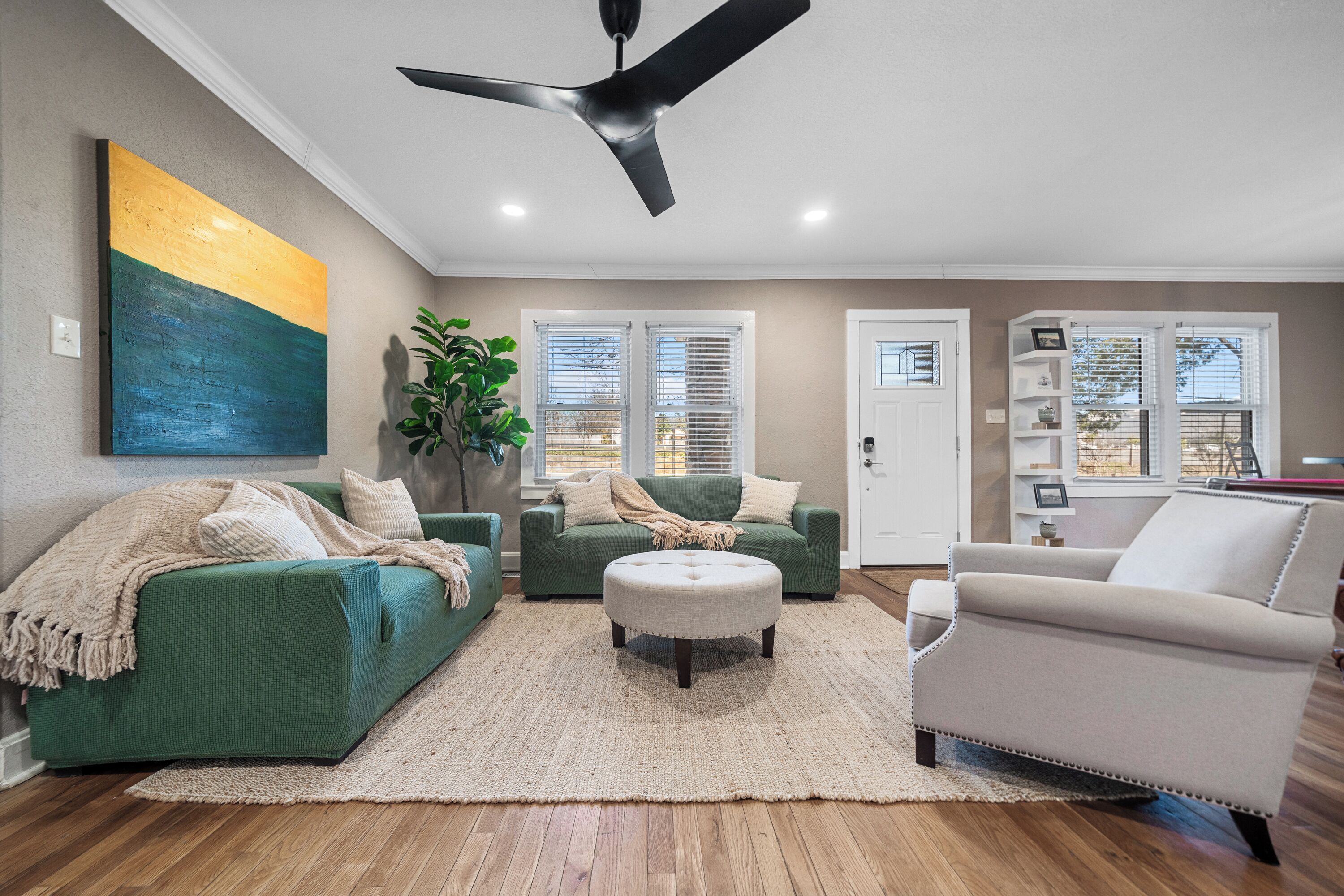 A living room with a green sofa, a white armchair, a beige ottoman, and a ceiling fan.