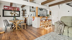 A spacious living area with a wooden dining table, bar stools, and a sofa. A kitchen is visible in the background with white cabinets and a wooden countertop. - Summit Slope Side + Steps 2 Slopes! (Big Bear Lake)