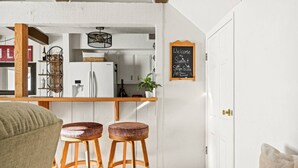 A cozy living area with a kitchen in the background, wooden stools, a chalkboard sign, and a white door. - Summit Slope Side + Steps 2 Slopes! (Big Bear Lake)