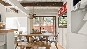 A dining area with a wooden table and chairs, a hanging light fixture, and a view of the outdoors through a large window. - Summit Slope Side + Steps 2 Slopes! (Big Bear Lake)