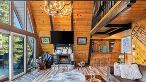 A cozy cabin interior with a stone fireplace, wooden floors, a leather sofa, and a large window overlooking the deck. - Luxury A-Frame at Bear Mtn | Hot tub and game room (Big Bear Lake)