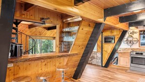 A rustic kitchen with wooden walls, a stone fireplace, and a bar area with stools. - Luxury A-Frame at Bear Mtn | Hot tub and game room (Big Bear Lake)