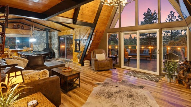 A cozy interior with a stone fireplace, wooden furniture, and a large glass door leading to an outdoor deck. - Luxury A-Frame at Bear Mtn | Hot tub and game room (Big Bear Lake)