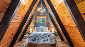 A cozy attic bedroom with a sloped ceiling, a bed with a fluffy blanket, a chandelier, and a window with a view of greenery. - Luxury A-Frame at Bear Mtn | Hot tub and game room (Big Bear Lake)