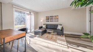 Una sala moderna con un sofá seccional, una mesa de comedor de madera y un ventanal con vistas a árboles.