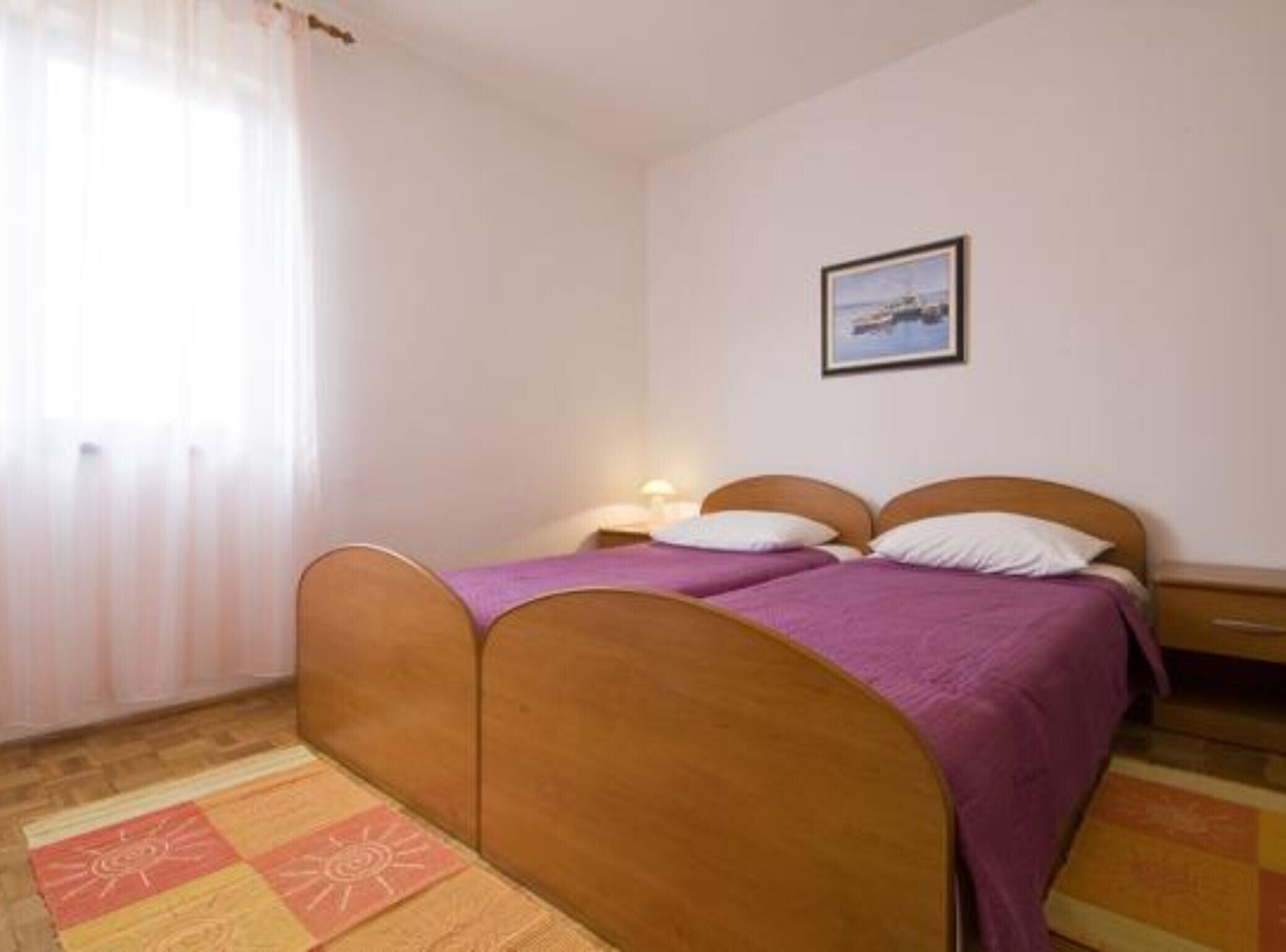 A bedroom with two single beds, a nightstand, a lamp, a framed picture on the wall, and a window with sheer curtains.