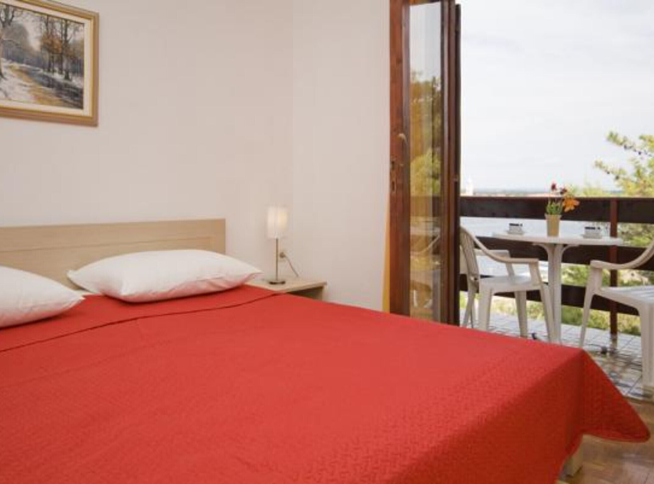 Une chambre d’hôtel avec un lit, une table de chevet avec une lampe, un tableau au mur et un balcon avec une table et des chaises.