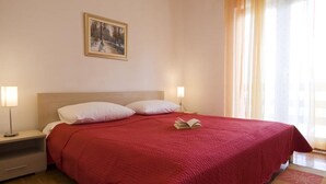 Una cama bien tendida con un edredón rojo, dos lámparas de noche, una mesita auxiliar pequeña y un cuadro en la pared.