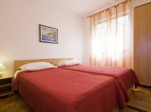 A room with two beds, a wooden headboard, a nightstand, a framed picture on the wall, and a window with sheer curtains.